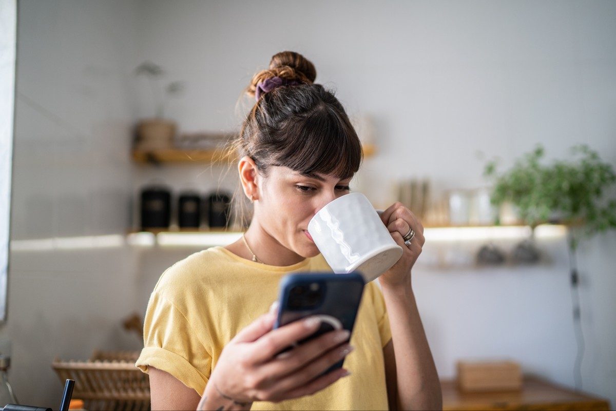Young woman using the mobile phone while drinking coffee or tea at home